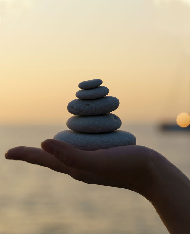 Hand holding a small stack of smooth dark stones against a soft sunset sky